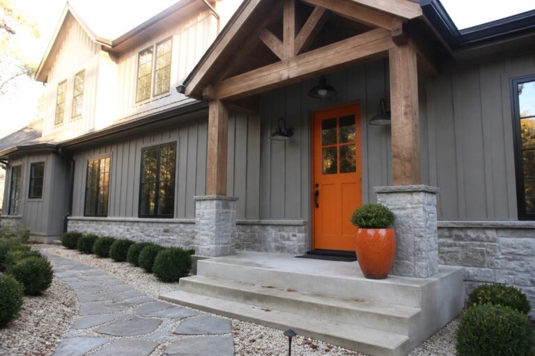 Front exterior of a gray house with an orange door, wooden beams, stone pillars, and a stone pathway leading to concrete steps.