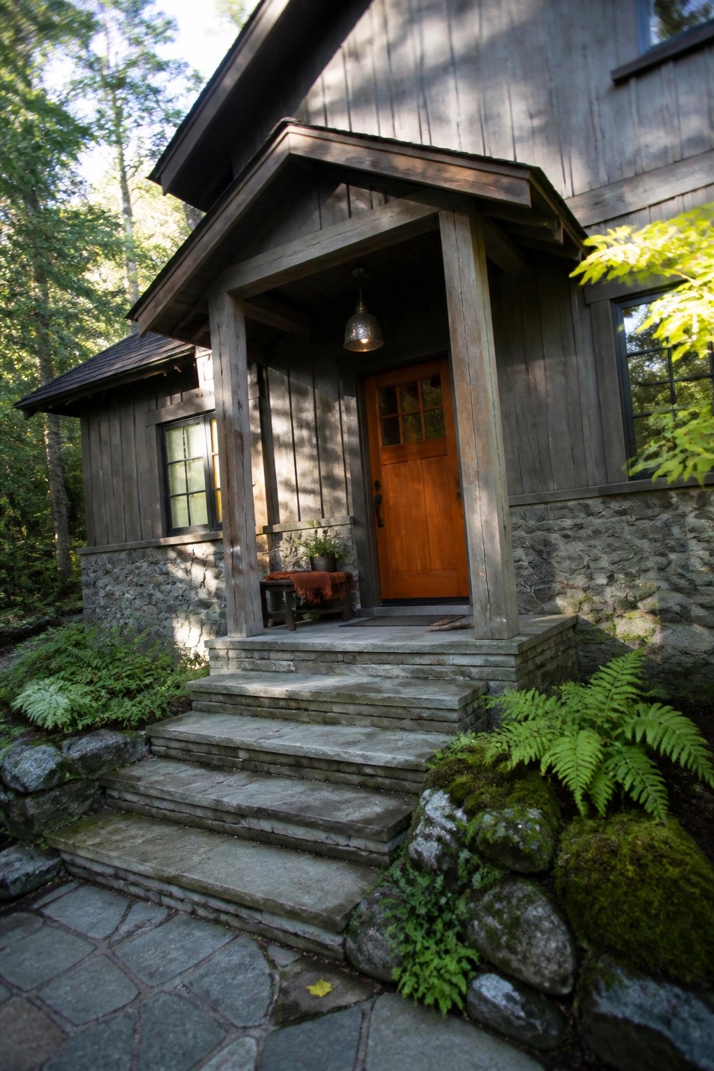 A gray wooden house with a stone foundation, an orange front door under a small porch, stone steps, and surrounding ferns and rocks.
