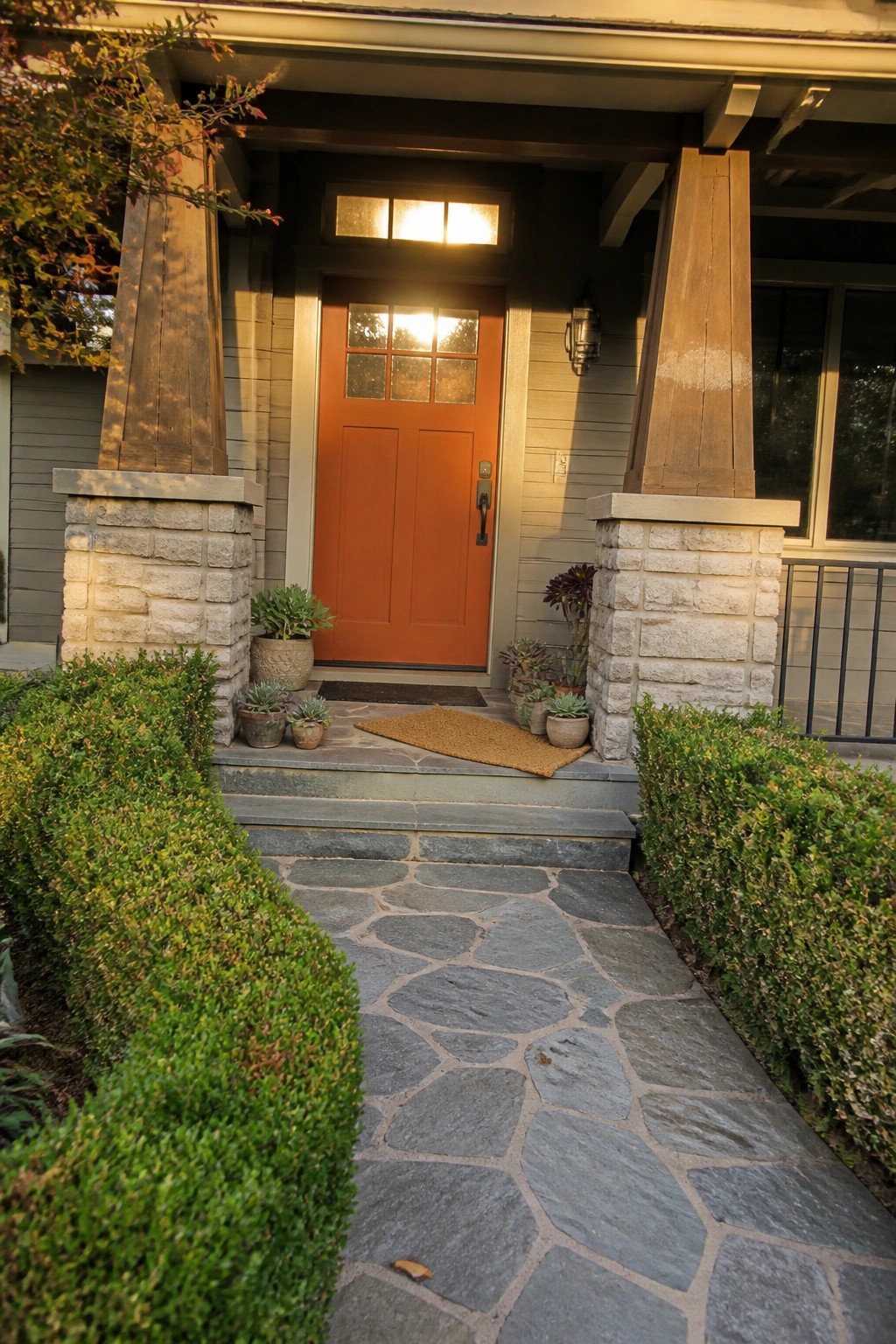 A house entrance with an orange door set between stone pillars, gray siding, and a stone pathway.
