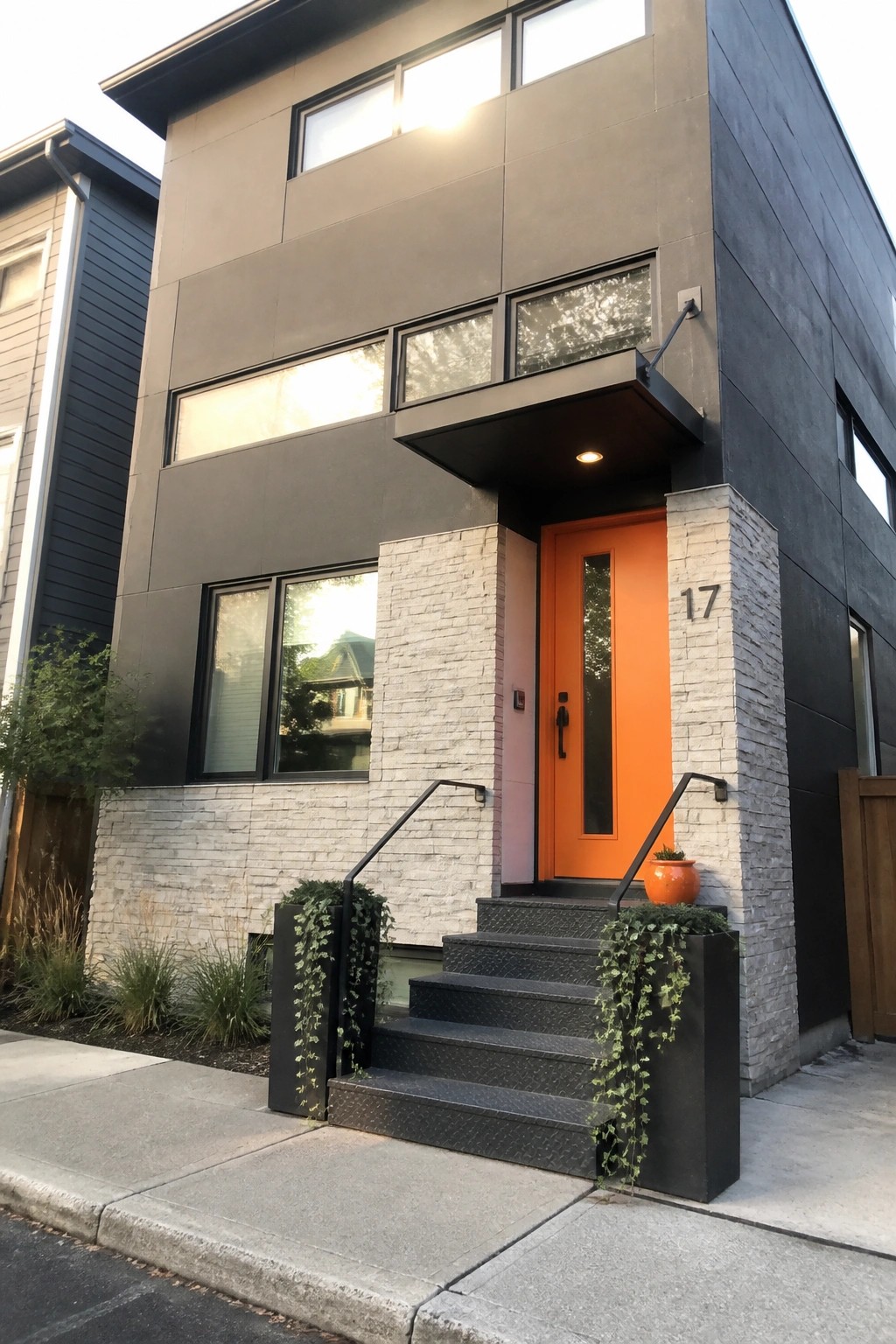 Modern two-story house with dark gray siding, bright orange front door, light stone pillars, and black metal stairs at the entrance.
