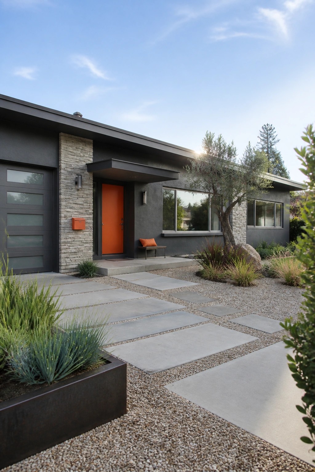 Modern single-story house with dark gray siding, light stone accents, bright orange front door, and concrete pavers set in gravel.