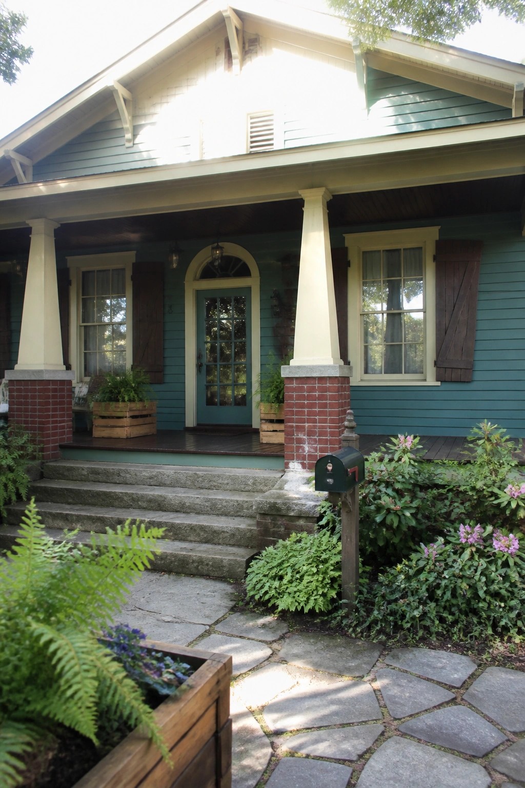 Front view of a teal Craftsman house with a covered porch, tapered white columns on red brick piers, a blue door, and stone steps leading to a flagstone path.