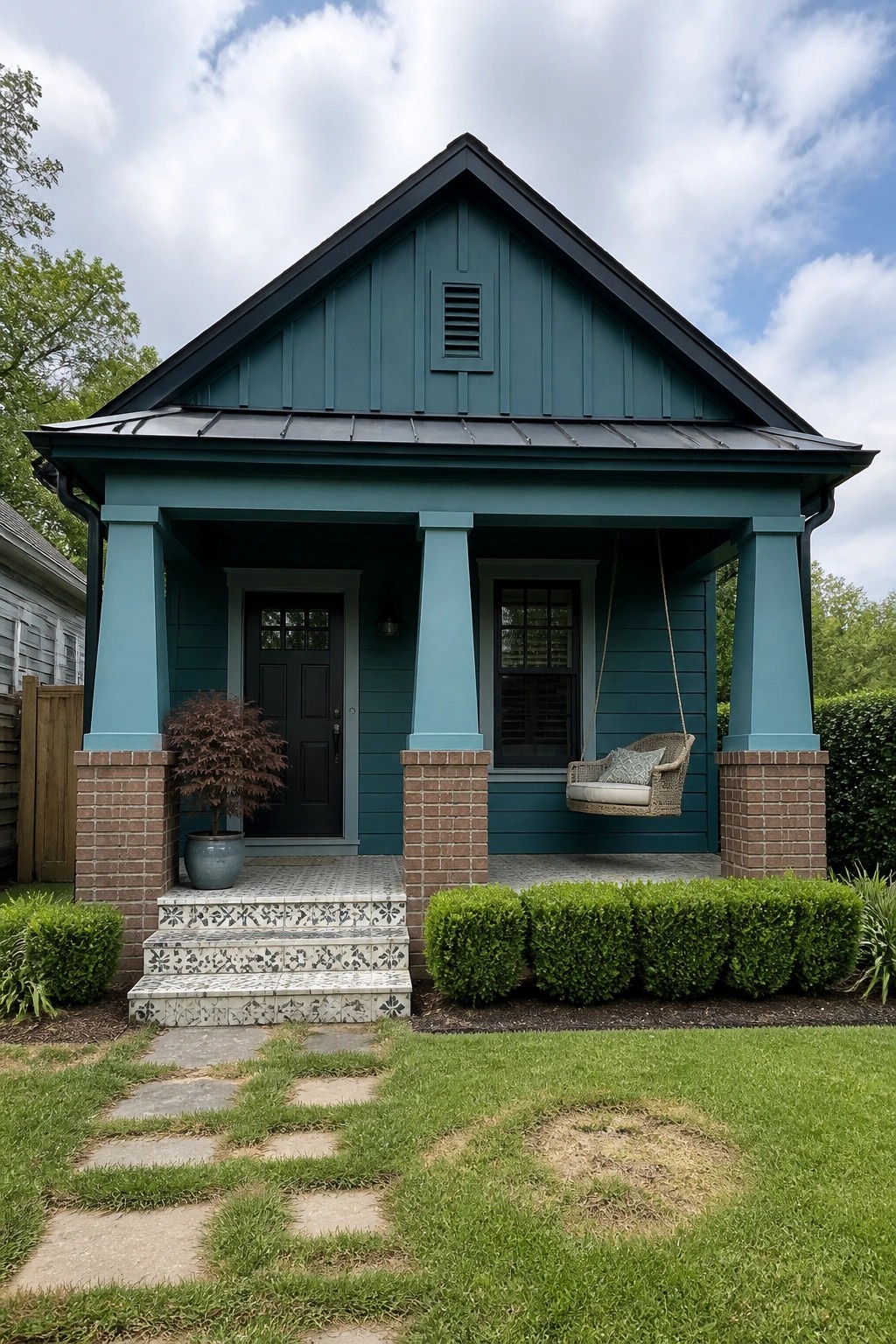 A teal Craftsman house exterior with a covered porch supported by tapered teal columns on brick piers, a hanging porch swing, patterned tile steps, and a small front lawn with stone path.