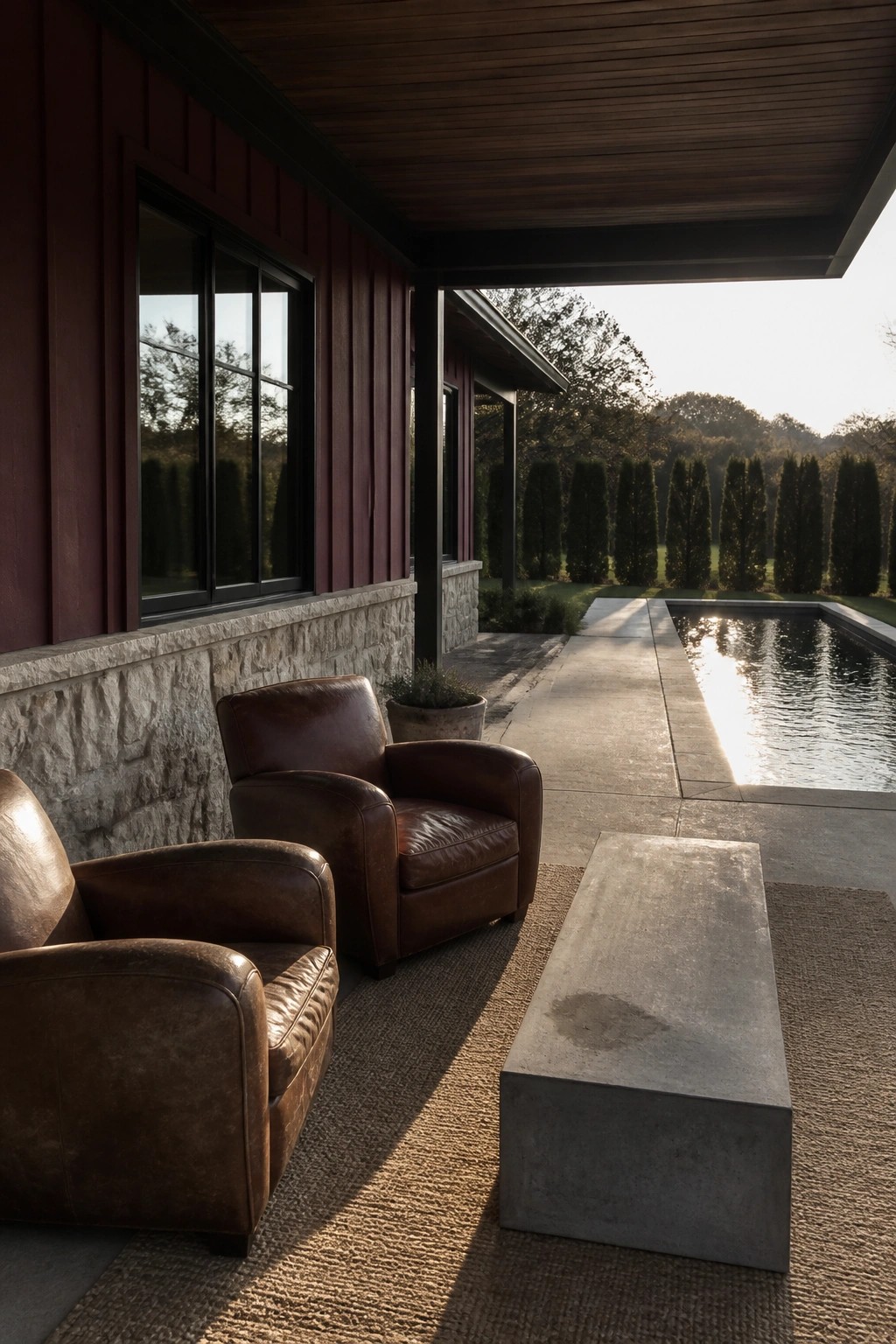 Two brown leather armchairs and a concrete bench rest on a woven rug beneath the covered porch of a plum-sided house with a stone foundation, overlooking a swimming pool.