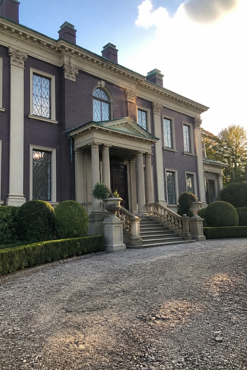 Grand purple brick mansion with white columns, stone steps, and gravel driveway with hedges