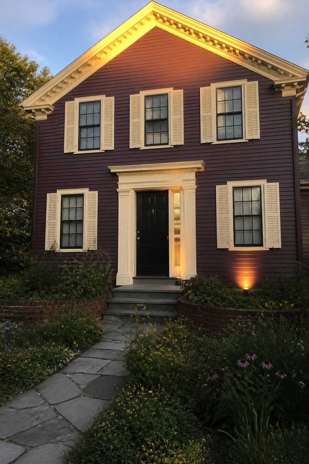 A two-story plum house with cream trim, white shutters, and a black front door at dusk, shown from the front with a stone path and garden beds.