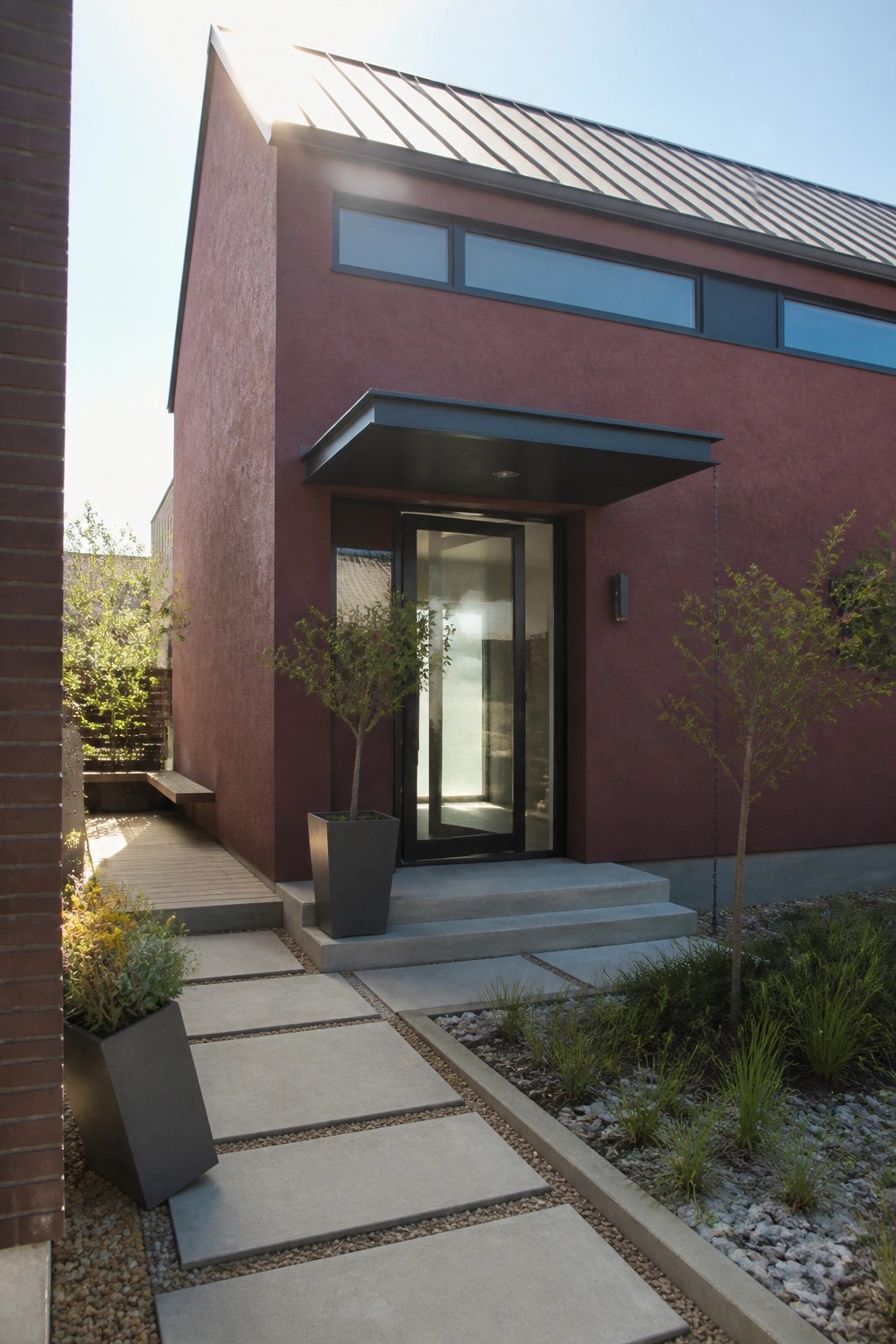 A maroon stucco house with a gray metal roof, black-framed glass door under a dark rectangular canopy, concrete steps, and a paver pathway leading to the entrance.