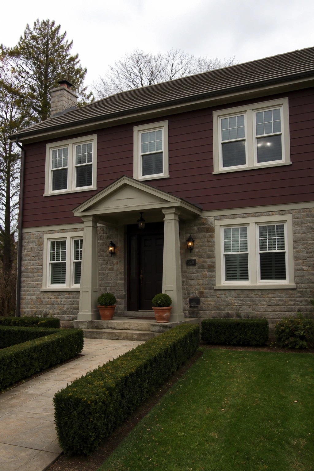 Two-story maroon house with stone base, columned porch, white windows, and green hedges.