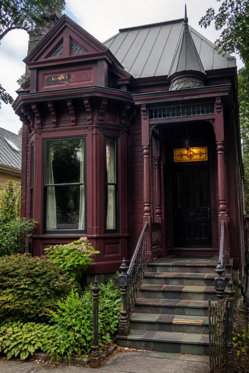 Ornate maroon Victorian house with bay window, black door, and stone steps