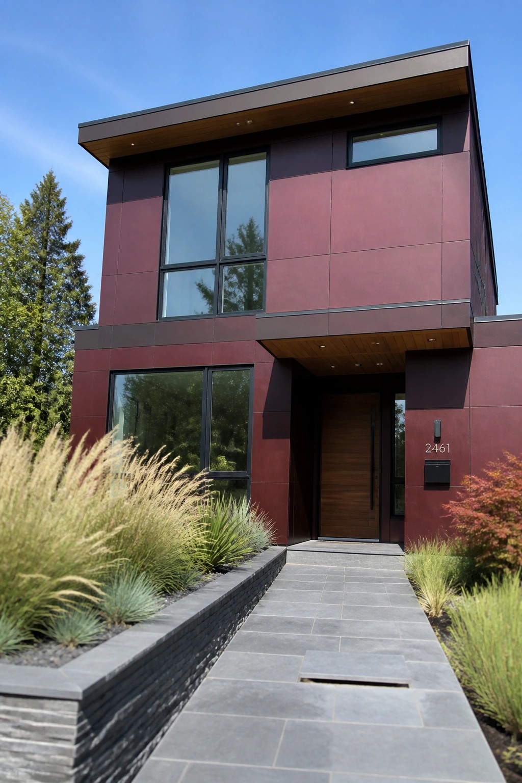 A two-story modern house with maroon panel siding, large black-framed windows, a wooden front door under a covered entry with wood ceiling, and a gray stone walkway bordered by tall grasses.