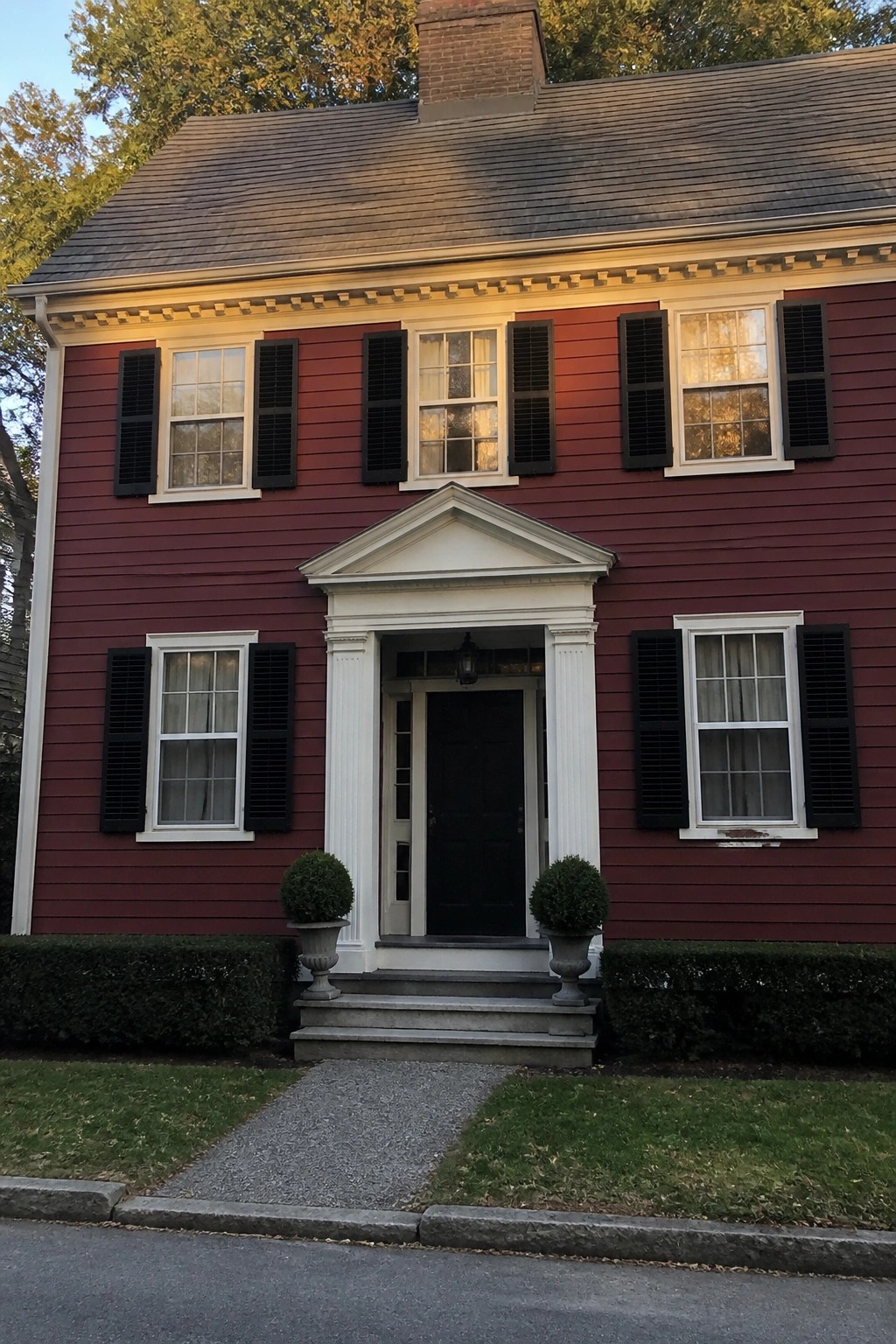 Red colonial house with black shutters, white portico, steps, and potted plants.