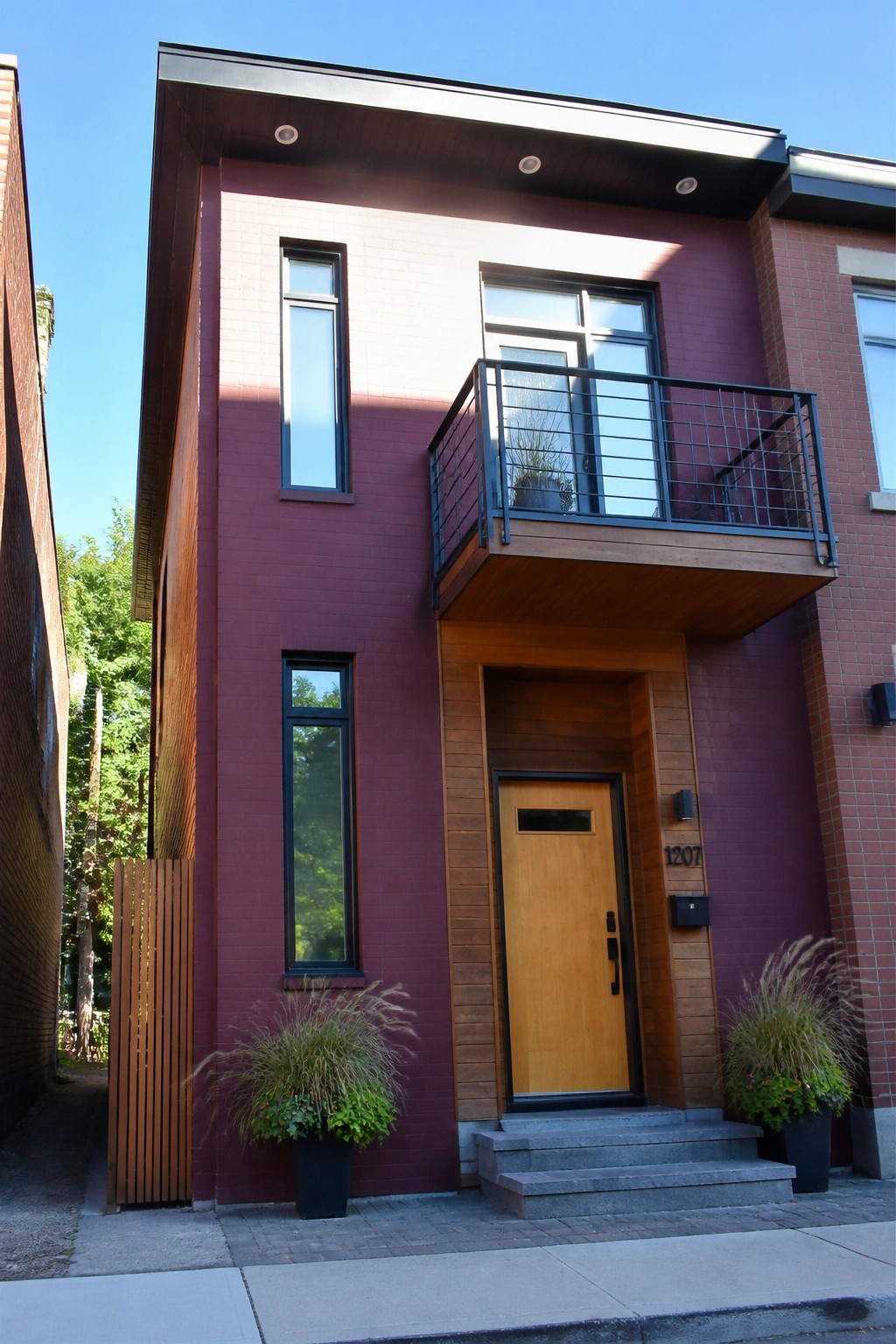 A two-story modern house with burgundy brick siding, a wooden front door and balcony structure, concrete steps, and potted plants beside the entrance.