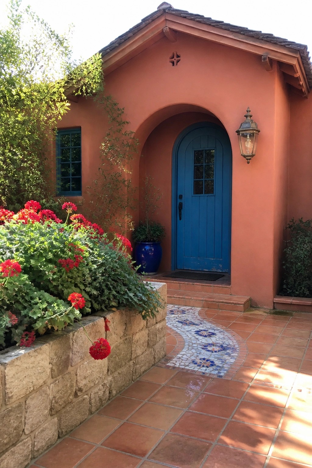 A terracotta stucco house exterior with a blue arched wooden door, small window, and red geraniums along a low stone wall.