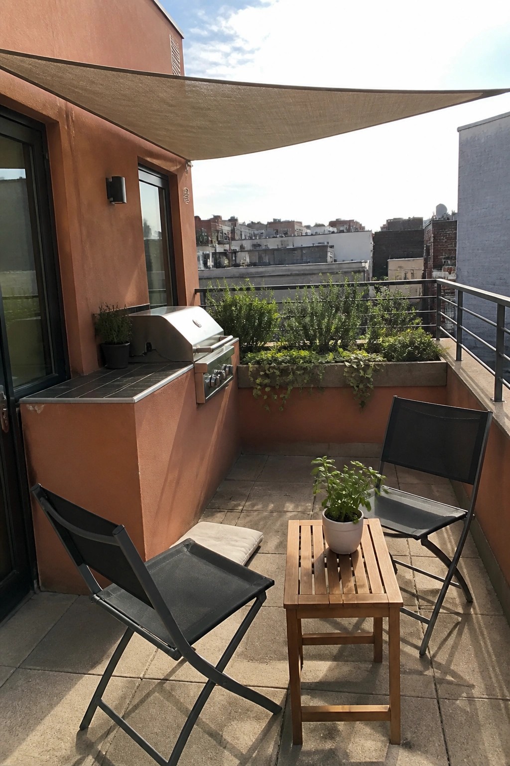 A compact balcony enclosed by terracotta stucco walls features a built-in grill, two black folding chairs, a small wooden table with a potted plant, and a beige shade sail overhead.