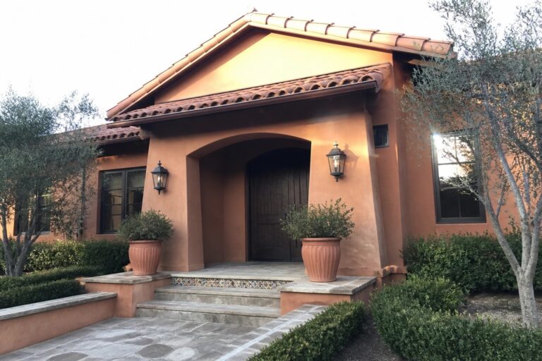 A terracotta stucco house with a recessed arched entry, wooden door, wall lanterns, and terracotta pots on the steps.