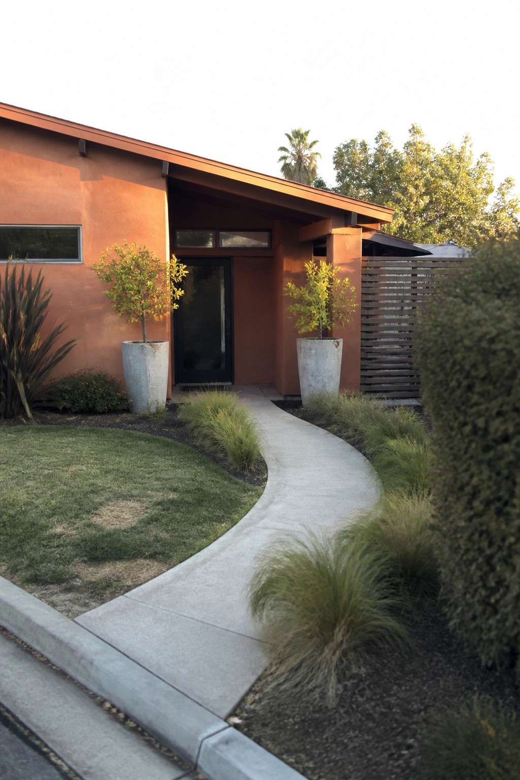 A house with smooth terracotta stucco walls, a dark entry door, and a curved concrete path bordered by grasses and large planters.