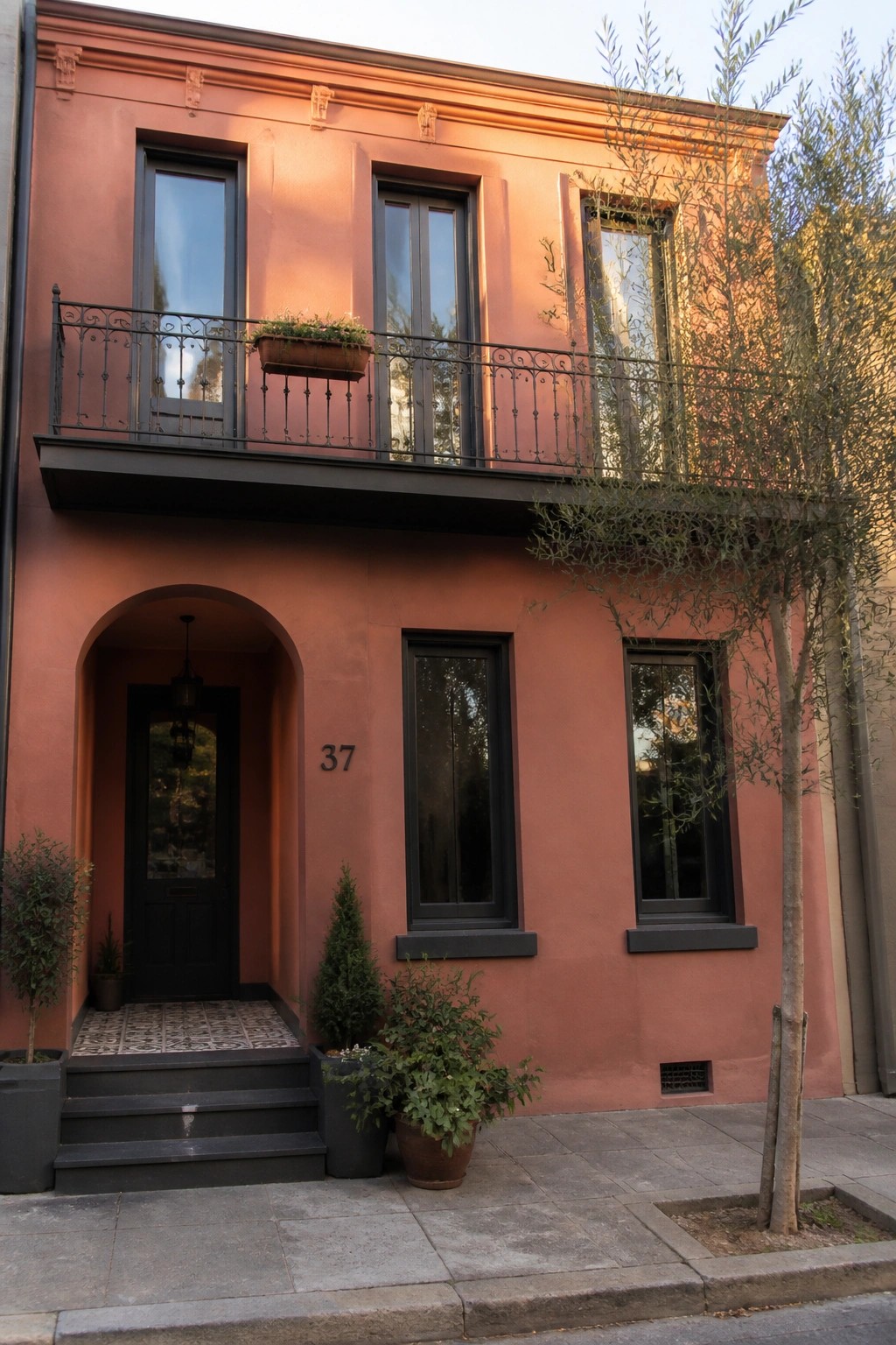 A two-story house with smooth terracotta stucco walls, dark-framed windows, a black metal balcony, and an arched entryway with a dark door.