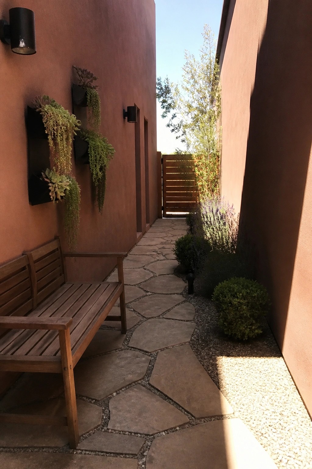 A narrow outdoor walkway lined with terracotta stucco walls, featuring a wooden bench on the left and multiple black wall-mounted planters holding succulents and trailing plants.