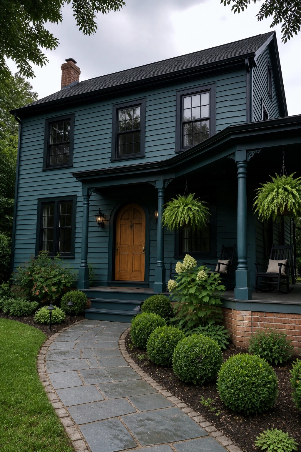A two-story house with dark teal siding, black window frames, a wooden front door, and a covered porch with rocking chairs.