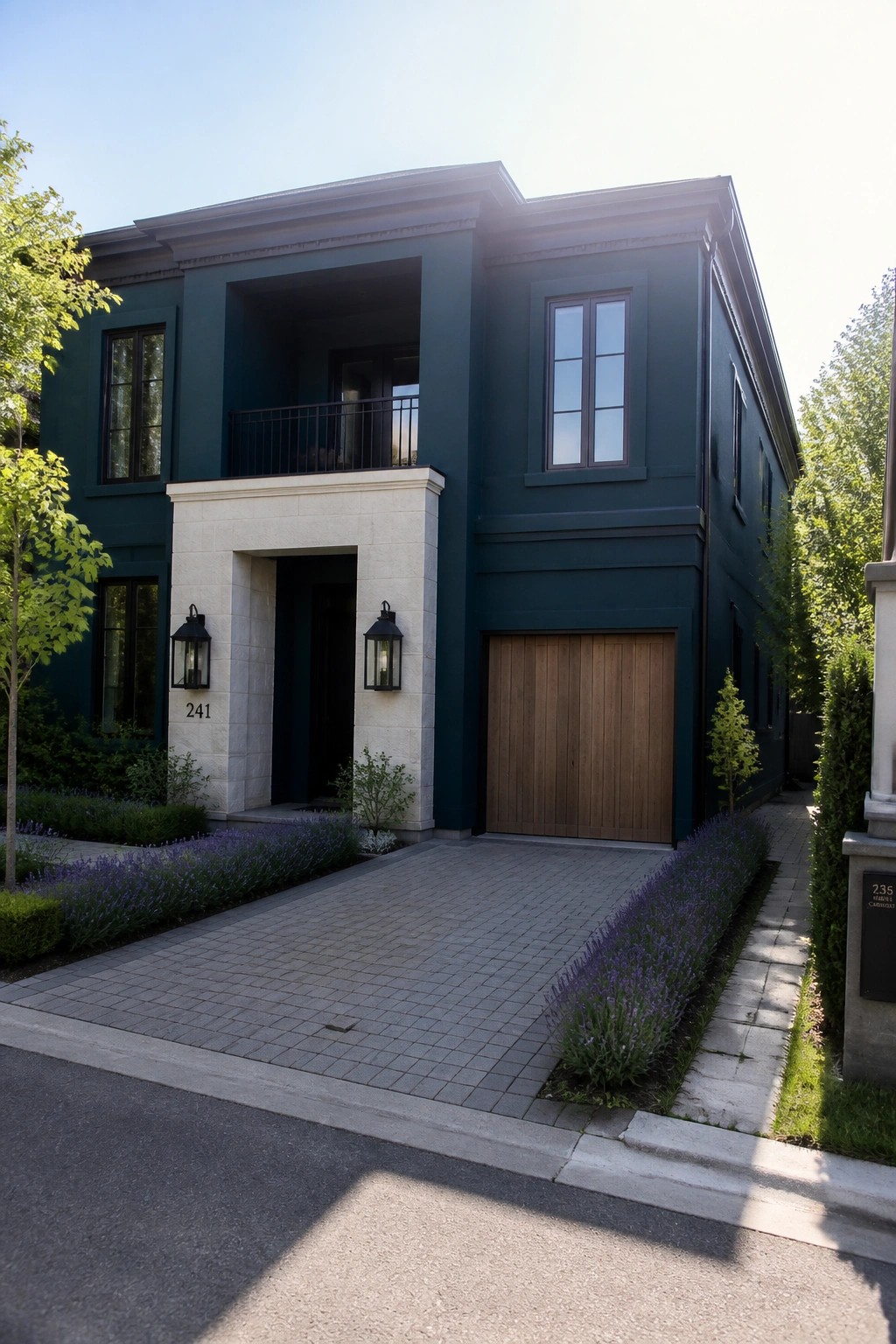 A two-story dark teal house with matte black windows, a stone entry surround, a wooden garage door, and purple flowering plants along the driveway.