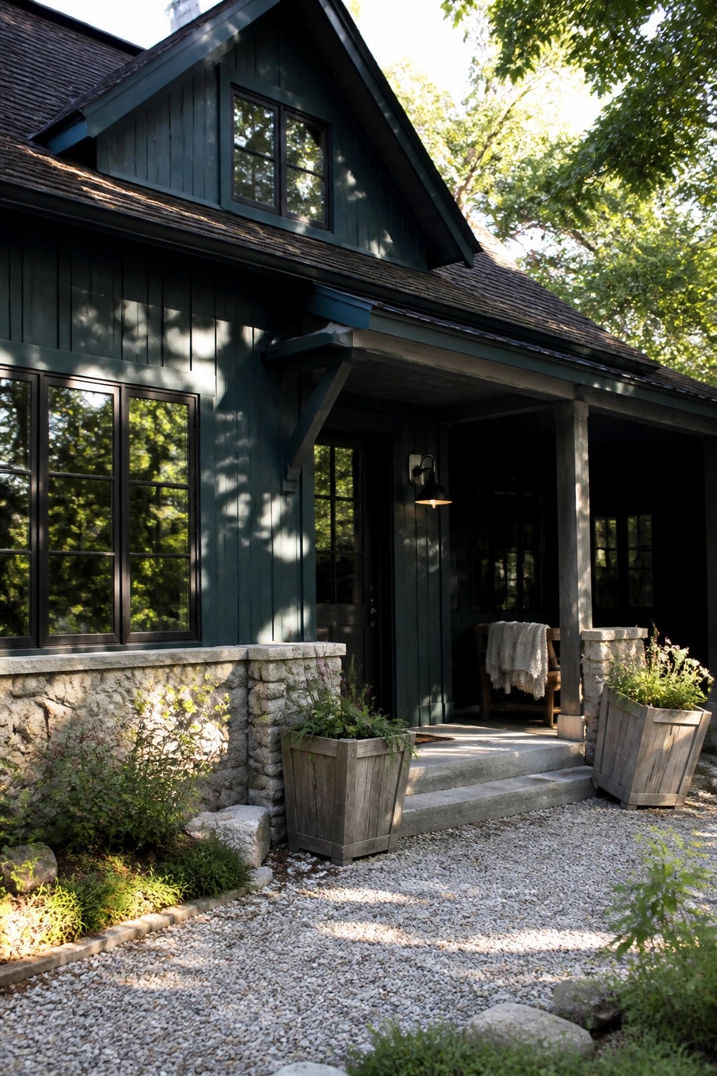 A dark teal house exterior with matte black windows, a stone foundation, gravel driveway, and a covered front porch.