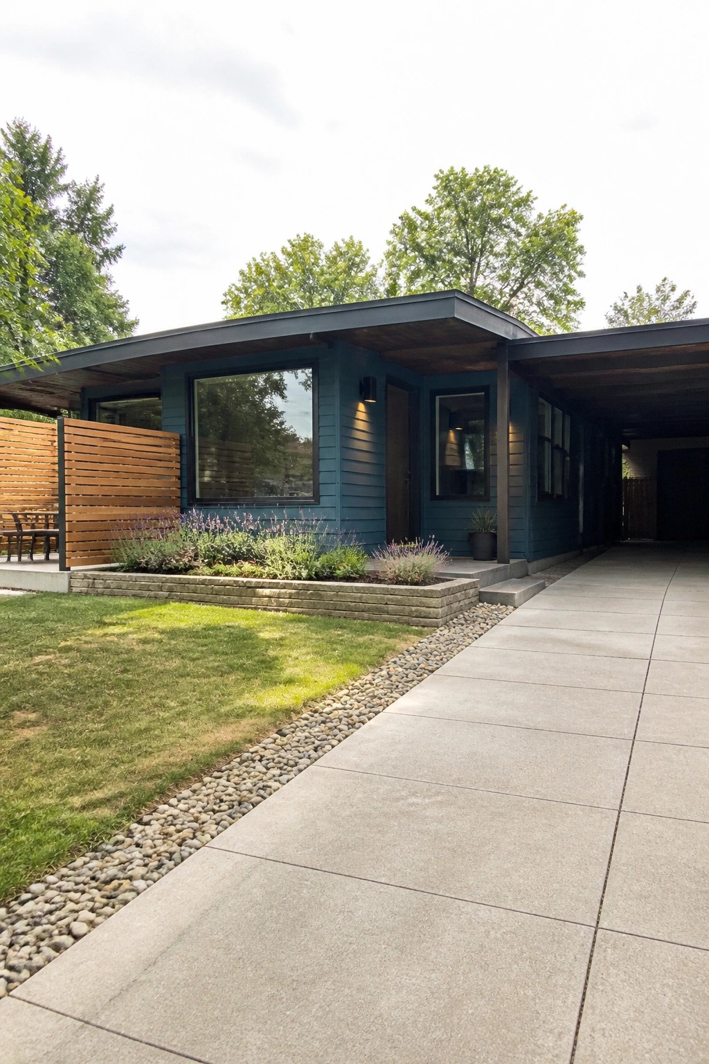 A single-story modern house with dark teal siding, matte black windows, a flat roof overhang, and a concrete driveway next to a gravel border with low plants.