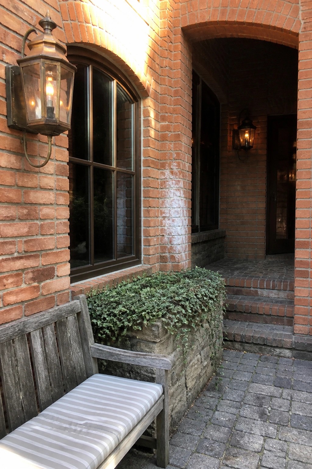 A close view of an orange brick house wall with a dark-framed arched window, bronze lantern, and stone steps leading to a doorway.