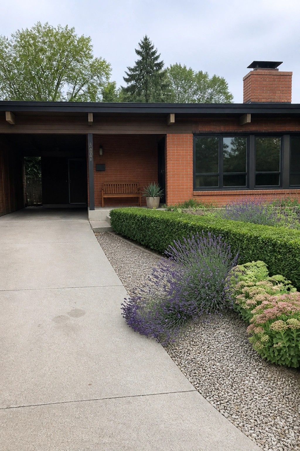 A single-story orange brick house exterior with dark bronze window frames, a covered carport on the left, and a gravel bed with lavender and green shrubs in the foreground.