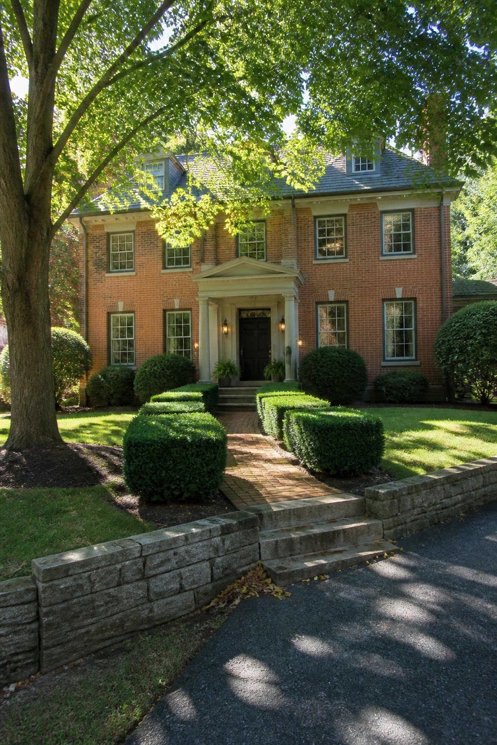 Red brick house with columned porch, manicured hedges, brick path, and stone wall.