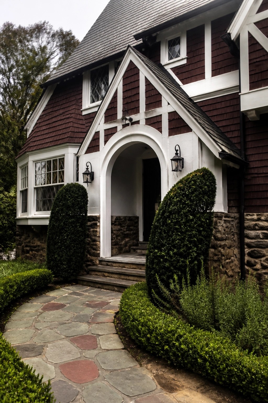 A maroon shingled house exterior with white trim featuring a prominent arched entryway, stone foundation, and flanking shrubs along a stone path.