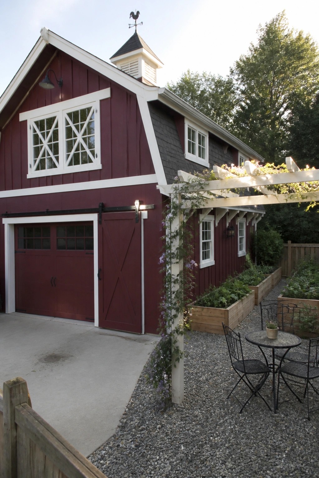 A two-story maroon house with white trim, black sliding barn doors, multiple windows, and a cupola with a weather vane, seen from a gravel side yard with raised garden beds and a small patio set.