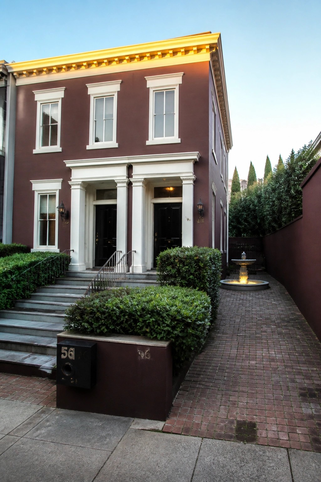 Maroon townhouse with white columns, gold cornice, brick path, and fountain.