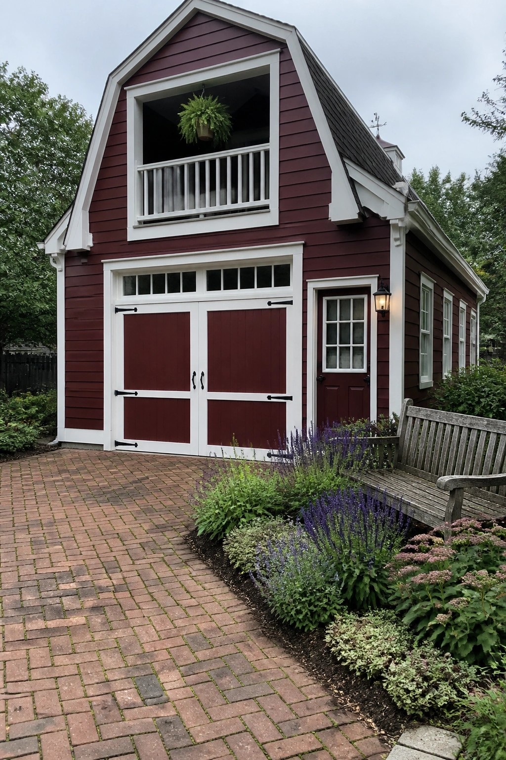 Red barn with white trim, garage doors, balcony, wooden bench, and flower garden.