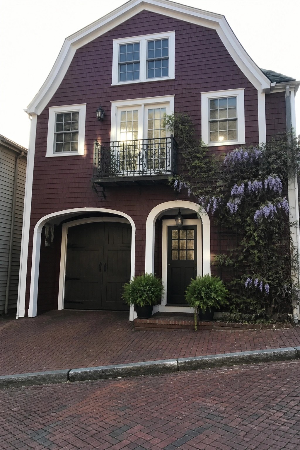 A two-story maroon shingle house with white trim shows an arched garage on the left, an arched front door on the right, and a small balcony above the garage.