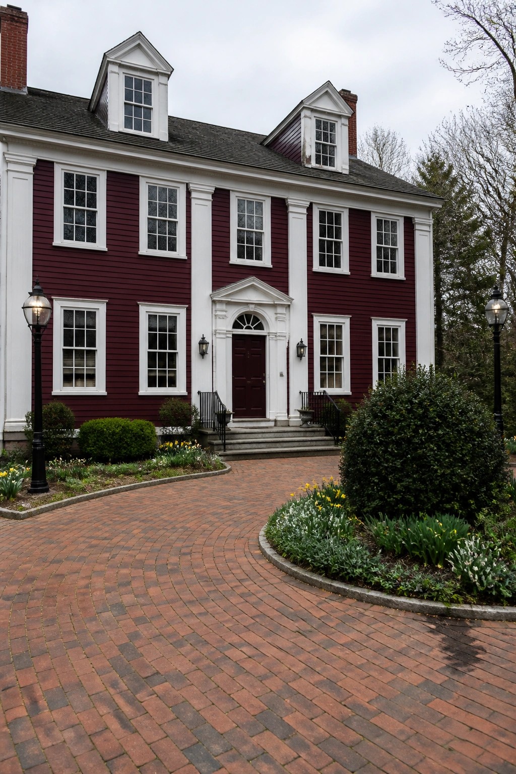 Red colonial house with white trim, brick driveway, and blooming gardens.