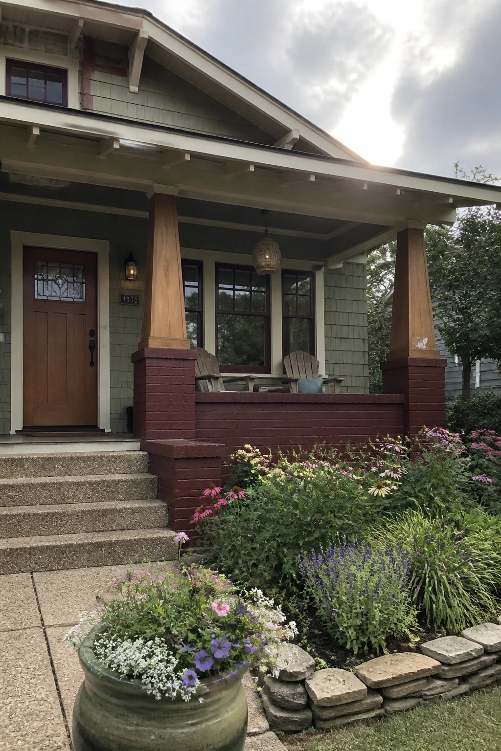Front view of a house with burgundy brick porch columns and base, green siding, wooden door, and flower garden along the front.