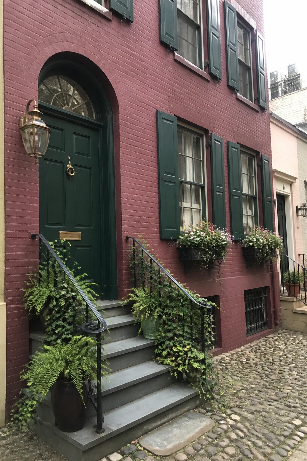 Red brick building with green door, shutters, stone steps, ferns, and flower boxes.
