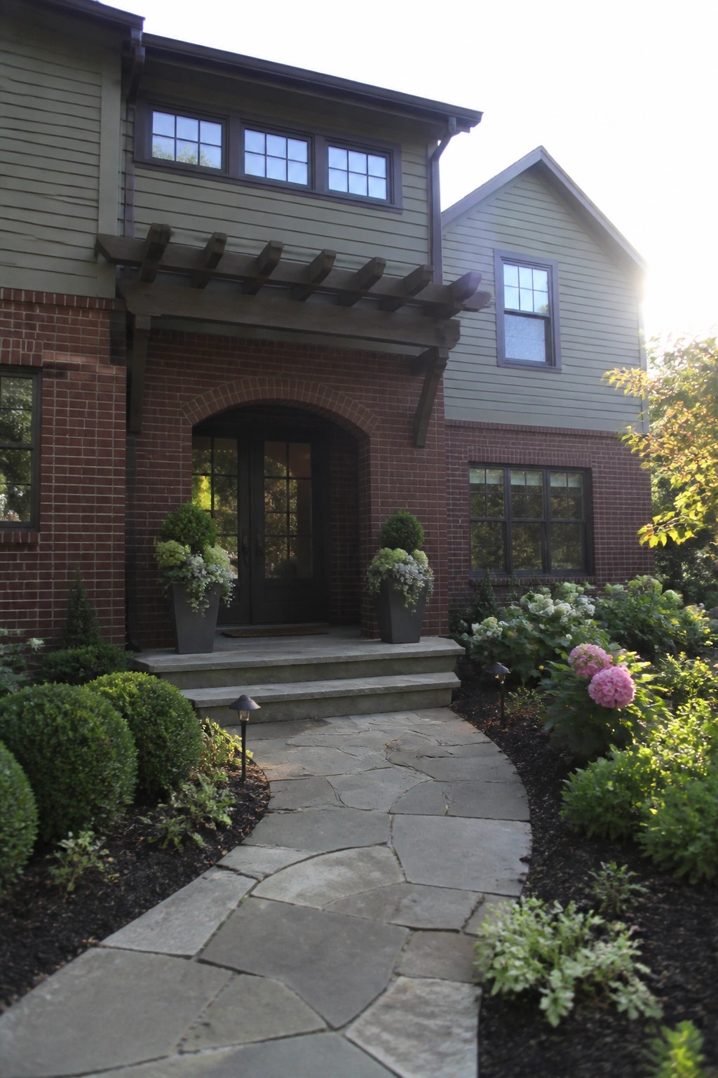 Stone pathway to arched brick entrance with potted plants and green siding.