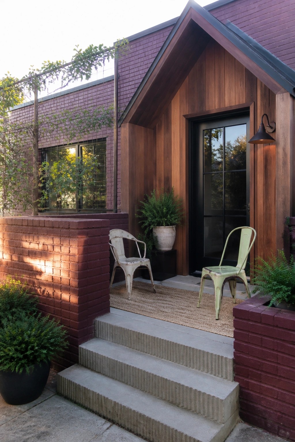 A burgundy brick house exterior features vertical wood siding around a black door, concrete steps, and two metal chairs on a woven mat.