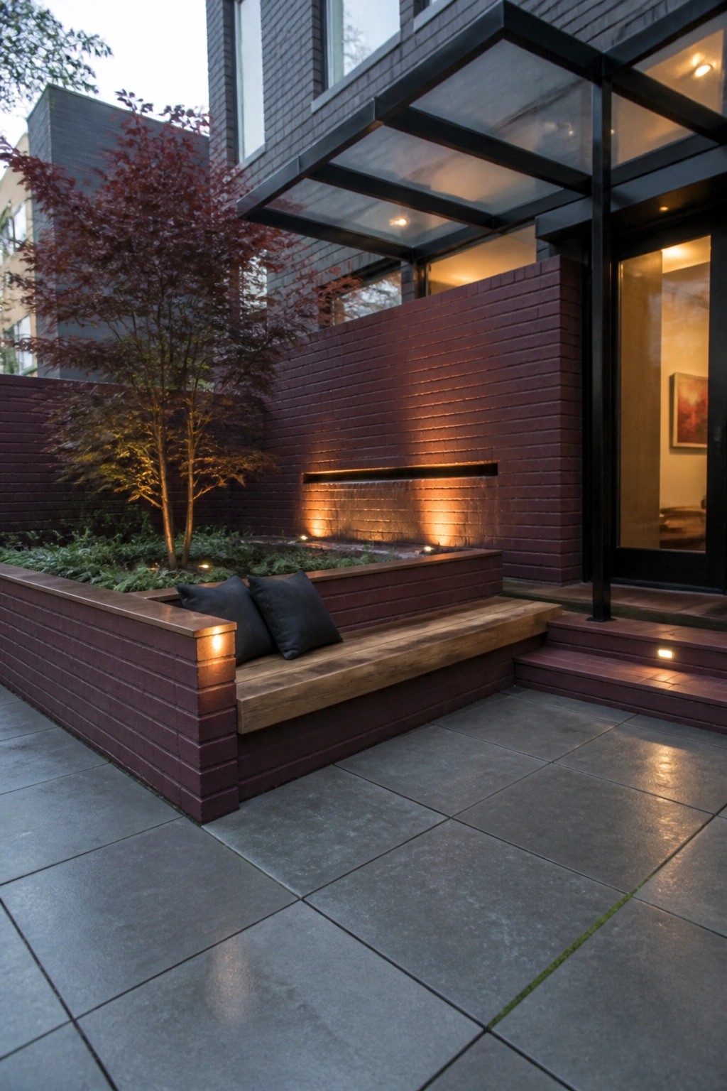 Modern brick patio with wooden bench, lit waterfall, and red maple tree
