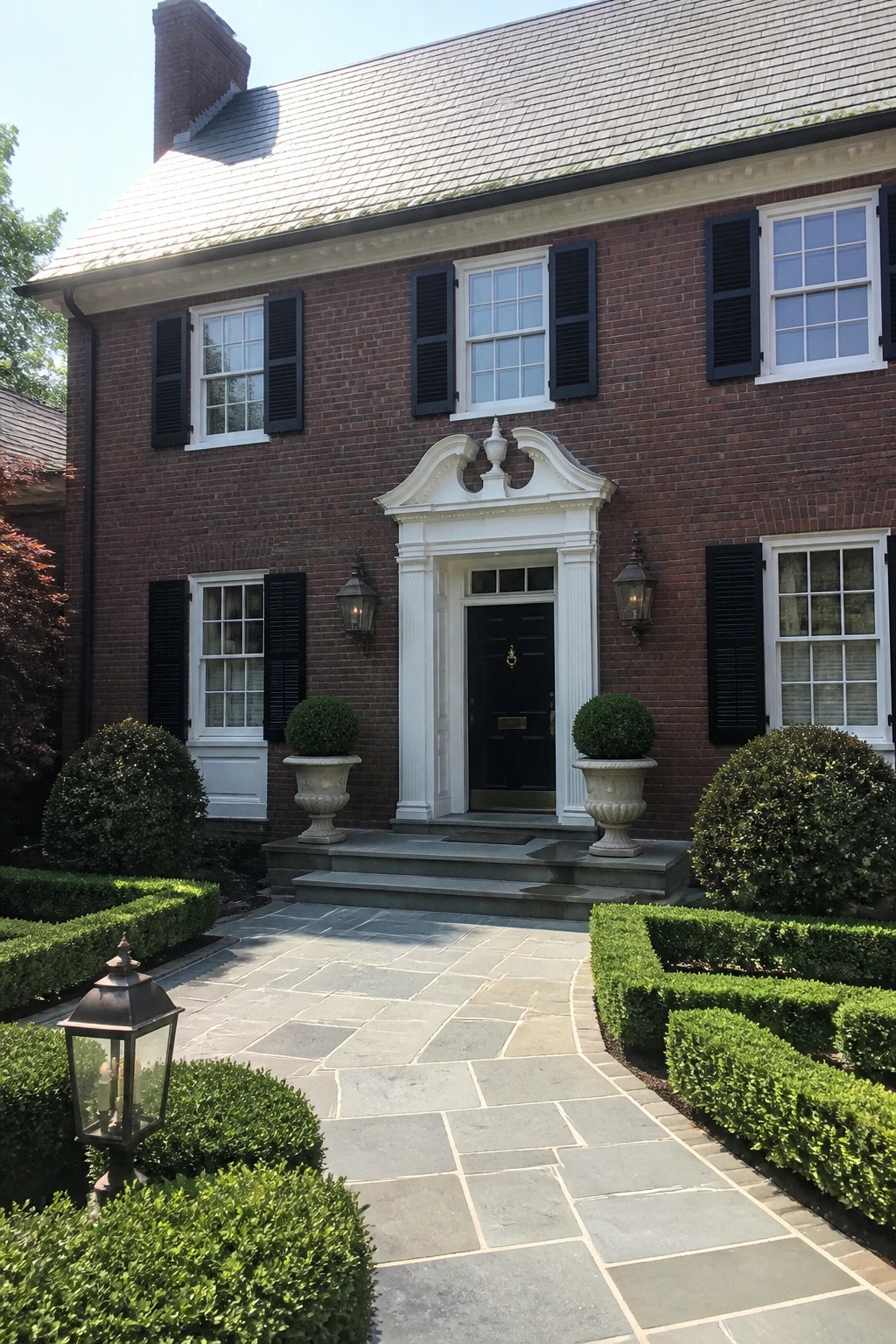 Red brick house with ornate white doorway, black shutters, stone path, and hedges.