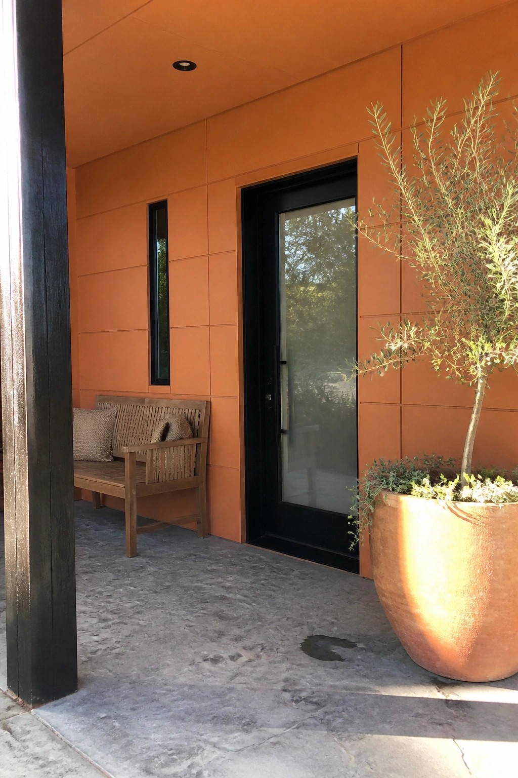 A house exterior clad in smooth orange fiber cement panels features a black door, a narrow vertical window, a wooden bench with cushions, and a large terracotta pot with a tree on a concrete surface.