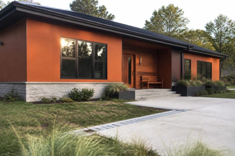 A modern single-story house with orange smooth fiber cement panels, a wooden entry porch, black-framed windows, and a stone foundation.