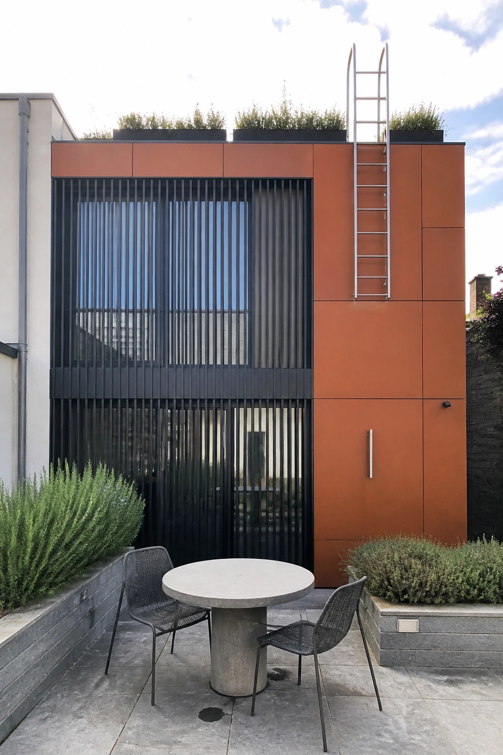 A modern house exterior clad in smooth orange fiber cement panels with black vertical slats over the windows and a concrete patio with round table and two chairs in front.