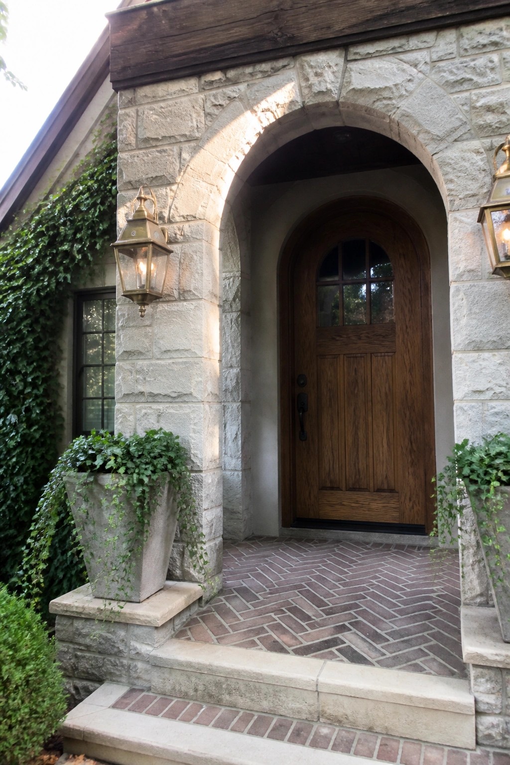 A wooden front door set inside a curved stone arch on a gray stone house, with lanterns on either side and brick pavers leading to the steps.