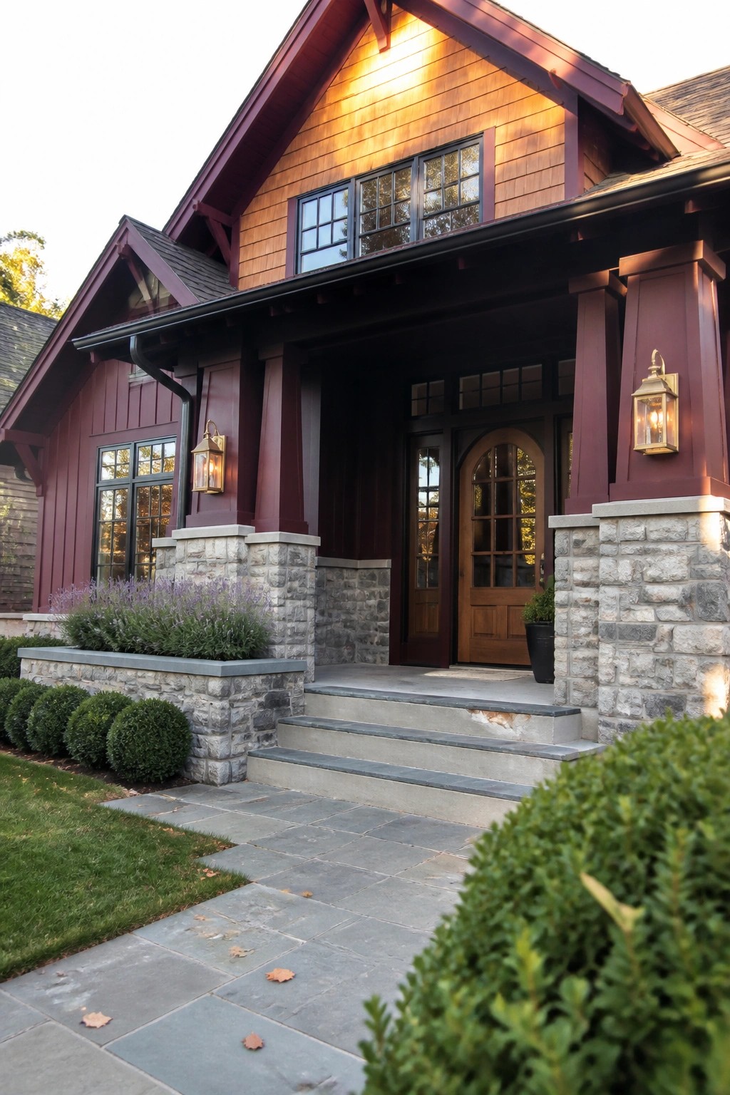 A maroon wood-sided house with gray stone pillars and foundation, a wooden front door, concrete steps, and green shrubs along a stone walkway.