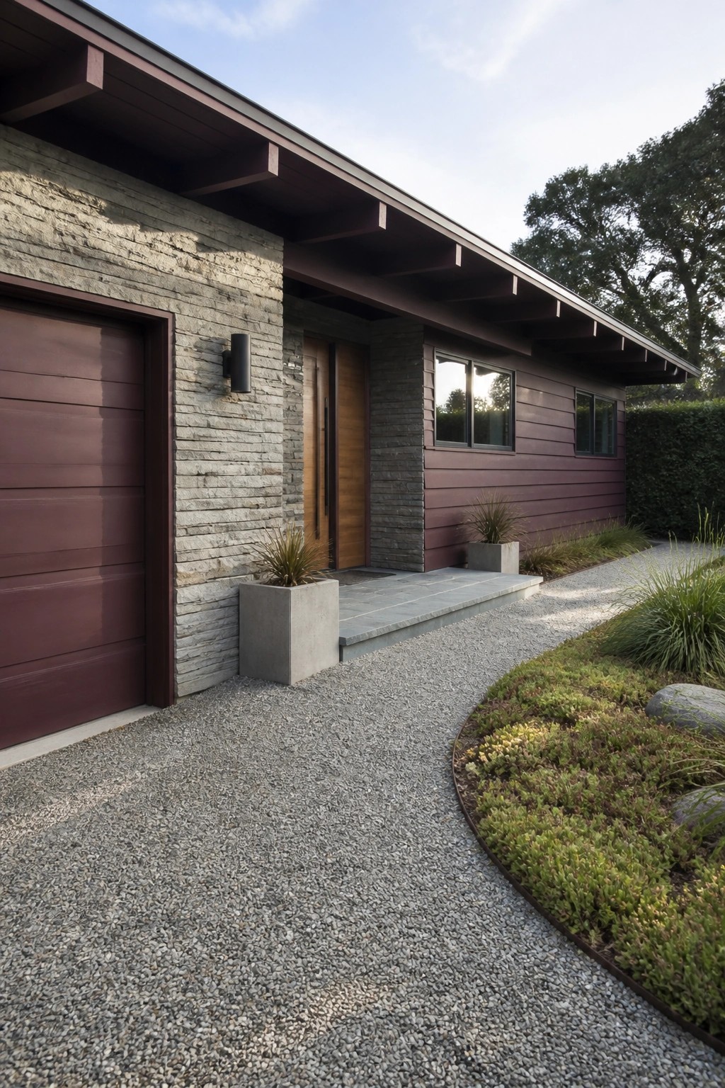 Modern single-story house exterior featuring maroon horizontal siding, gray stacked stone around the entry and garage, a wood front door, and a gravel driveway.