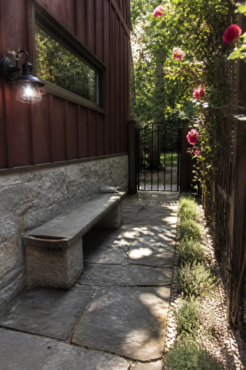 A burgundy wooden house exterior with a gray stone foundation, built-in stone bench, and stone pathway leading to a black metal gate.