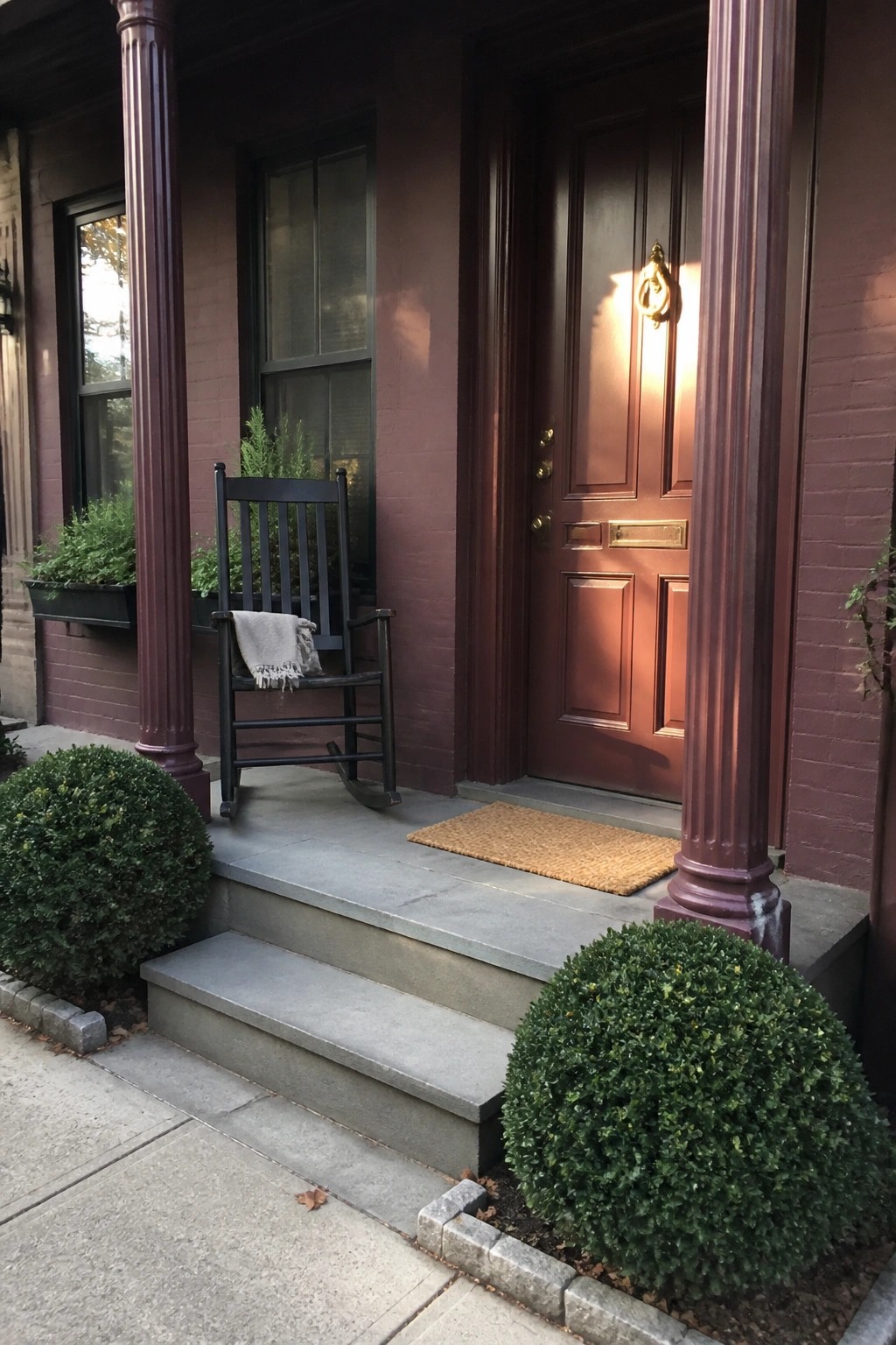 A burgundy house exterior with gray stone steps, a brown front door, black columns, and a black rocking chair on the porch.