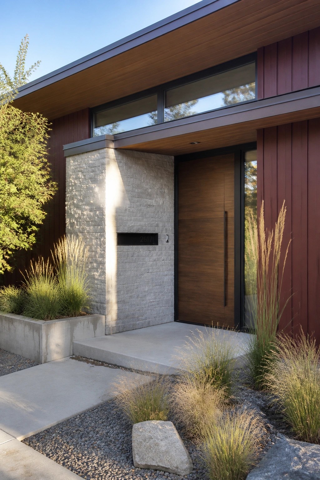 Modern house entrance with burgundy vertical siding, gray stone column, wooden door, and gravel landscaping with grasses.